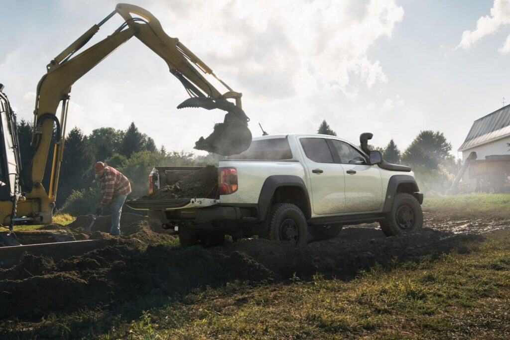 White Ford Ranger pickup truck with bed full of soil being loaded by excavator at rural property, worker standing nearby, forested background