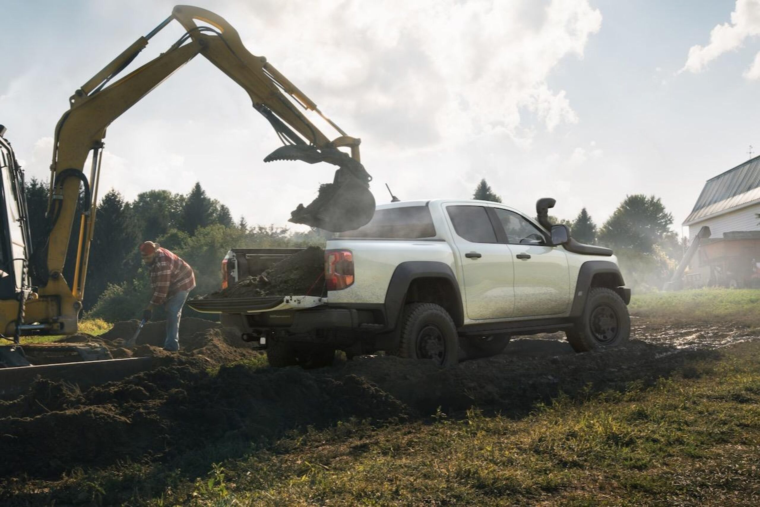 White Ford Ranger pickup truck with bed full of soil being loaded by excavator at rural property, worker standing nearby, forested background