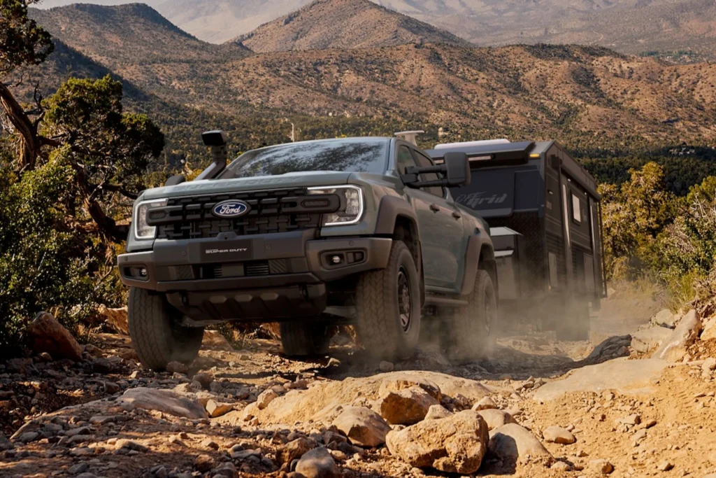 Ford Ranger Super Duty pickup truck towing a caravan on a rocky desert trail, kicking up dust, with mountains and scrubland in the background