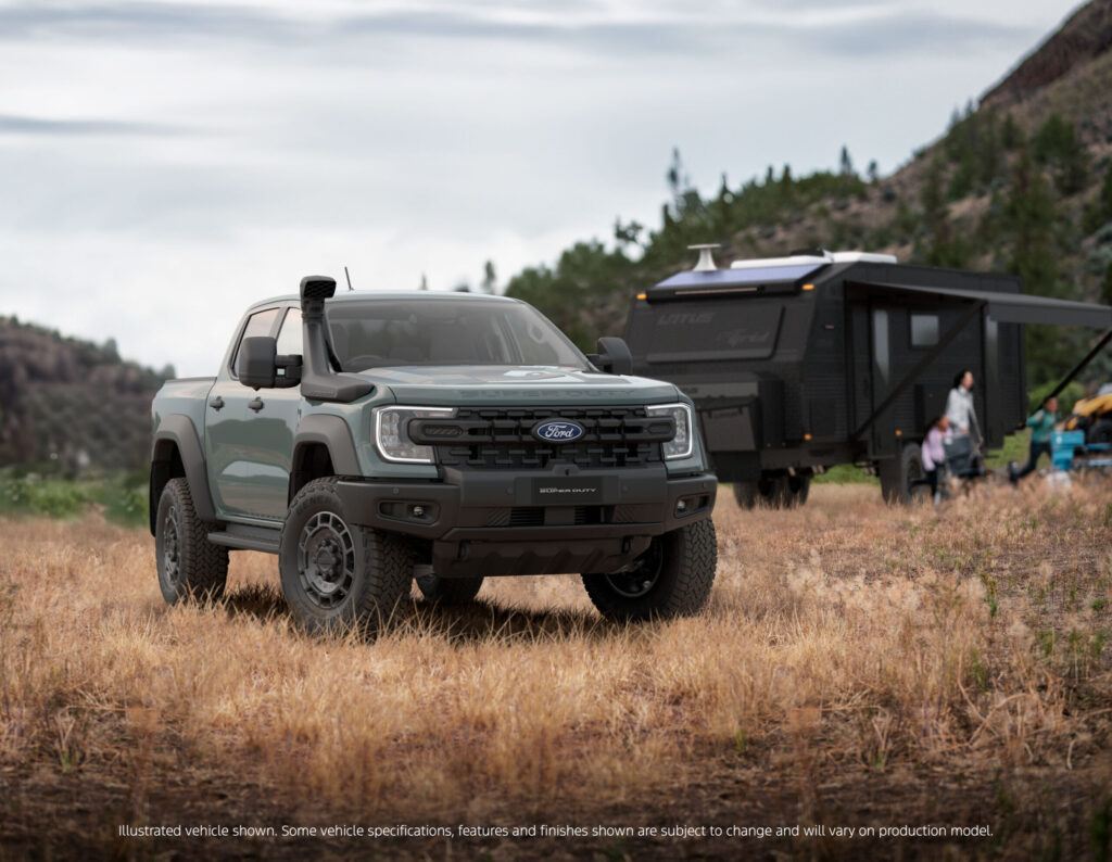 Silver Ford Ranger Superduty pickup truck parked in dry grassland with black travel trailer and people in background, mountains and evergreen trees visible