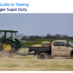Tan Ford Ranger Super Duty towing a green John Deere tractor with hay bales on a dirt road, motion blur effect