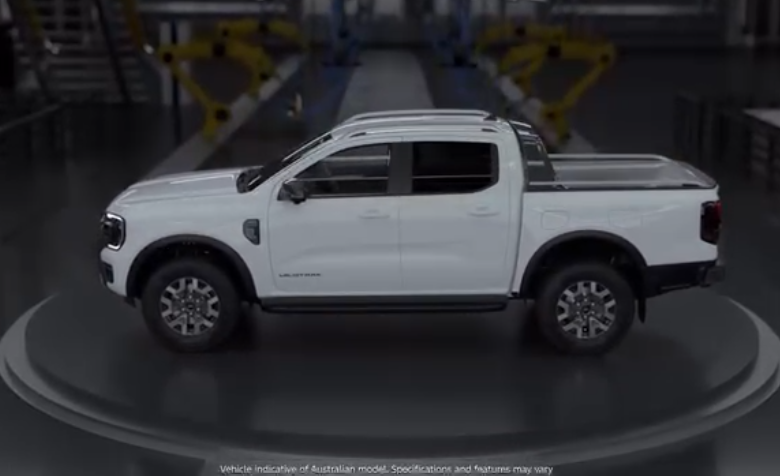 White Ford Ranger Hybrid pickup truck displayed on a rotating platform in an automotive showroom with industrial ceiling and yellow safety barriers visible in background