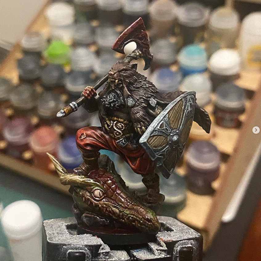 Painted fantasy miniature figurine of a skeletal warrior with dark armor, red cape, and large shield, standing on a dragon skull base, with paint bottles blurred in background