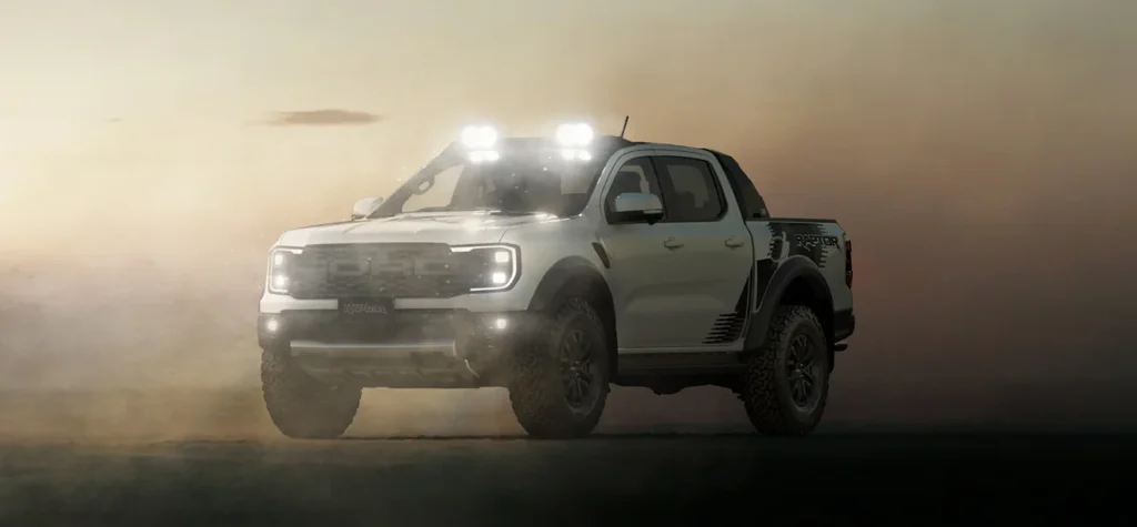 White Ford Ranger Raptor pickup truck with roof-mounted lights driving through dusty desert conditions with headlights on