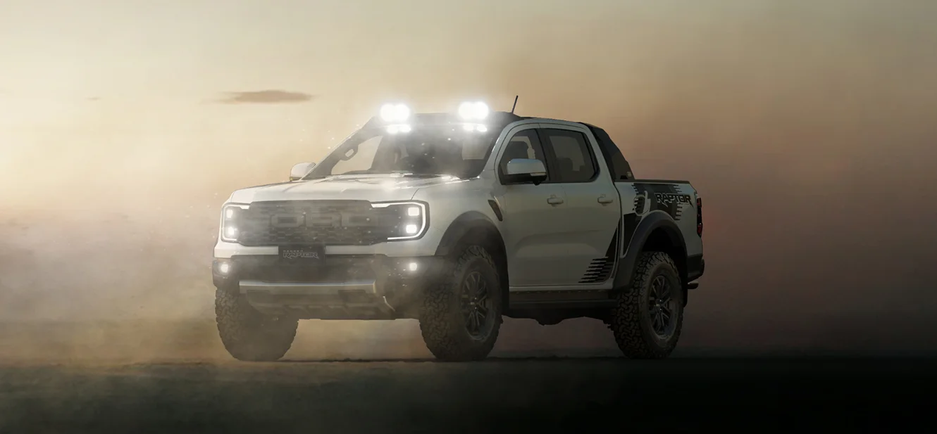 White Ford Ranger Raptor pickup truck with roof-mounted lights driving through dusty desert conditions with headlights on