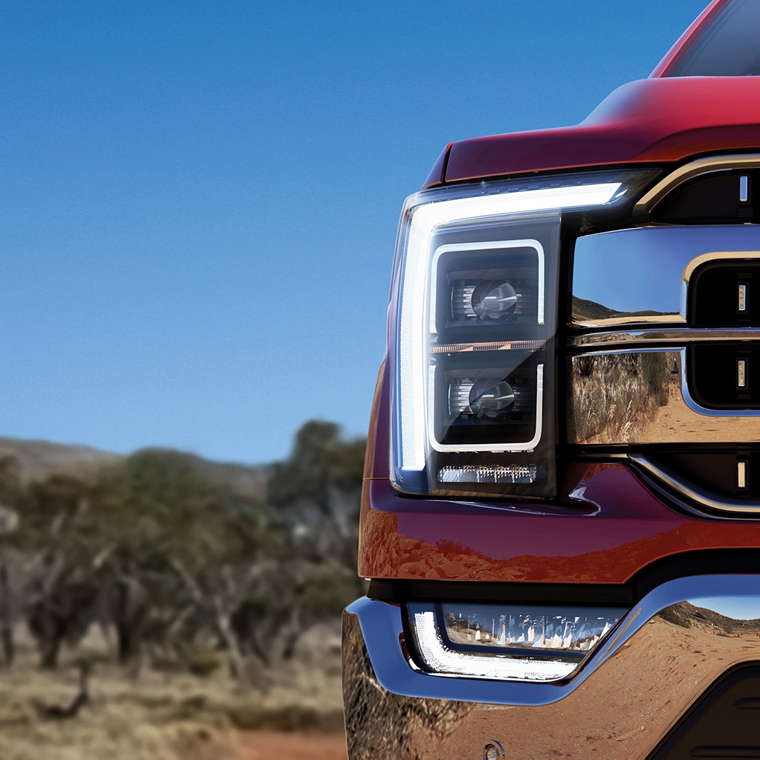 Close-up of a red Ford F150 truck's front end featuring LED headlamps with white accent lighting, parked in a desert landscape with clear blue sky