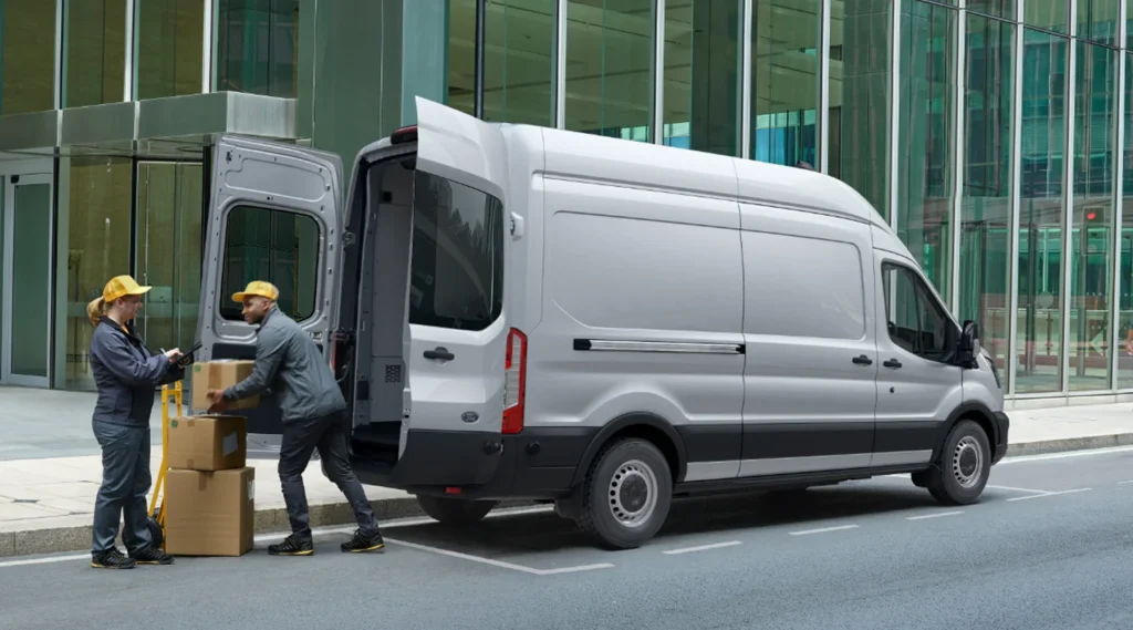Two workers in yellow hard hats loading cardboard boxes into a white commercial delivery van in front of a glass-fronted building