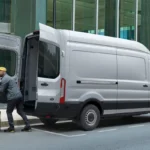 Two workers in yellow hard hats loading cardboard boxes into a white commercial delivery van in front of a glass-fronted building