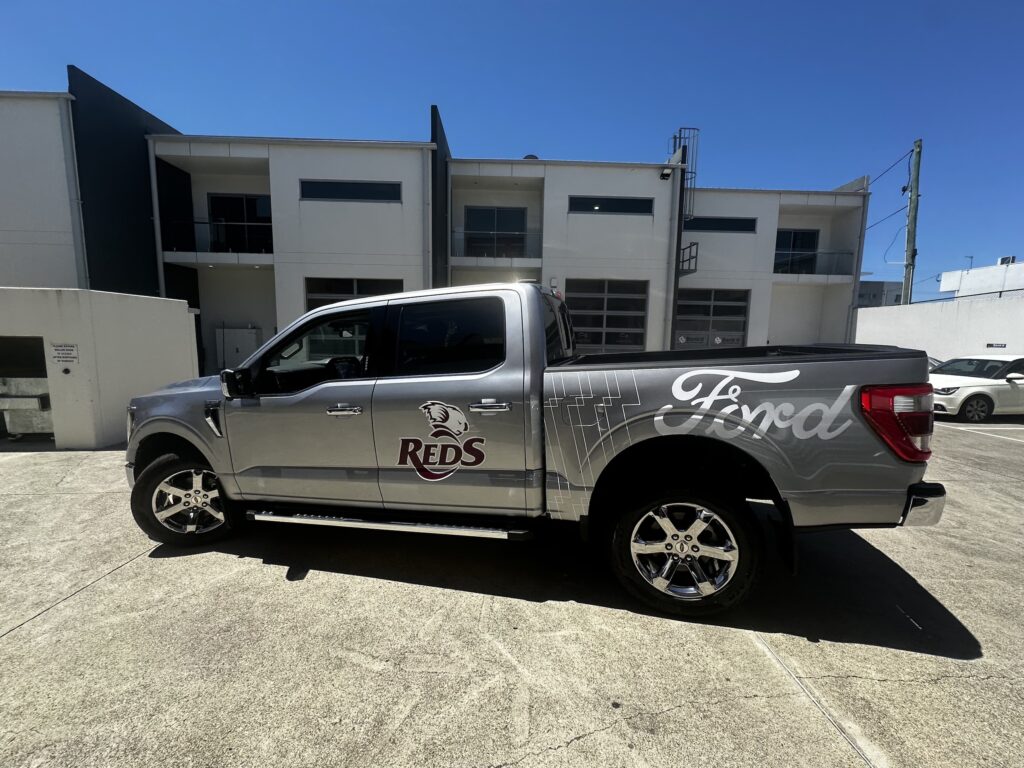 Silver Ford F150 pickup truck with Reds sports team livery parked in a concrete lot, featuring custom graphics on the bed and side panel, chrome wheels, modern industrial buildings visible in background