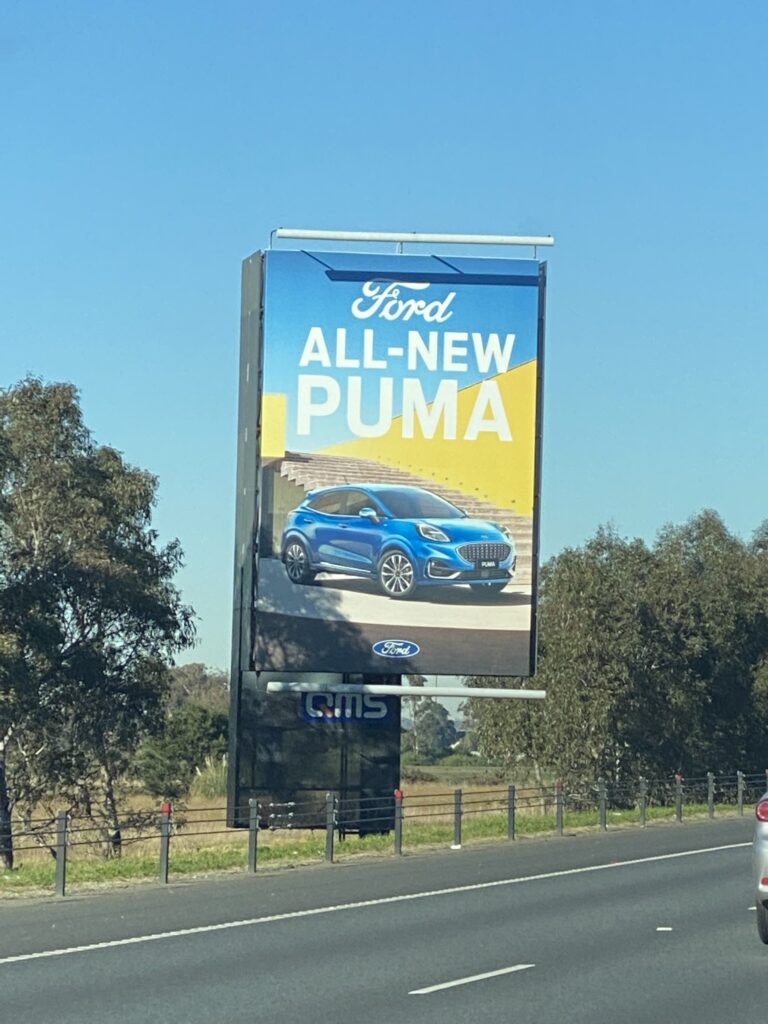 Large roadside billboard advertising Ford's all-new Puma with blue vehicle displayed against yellow and blue background, flanked by trees and rural fencing