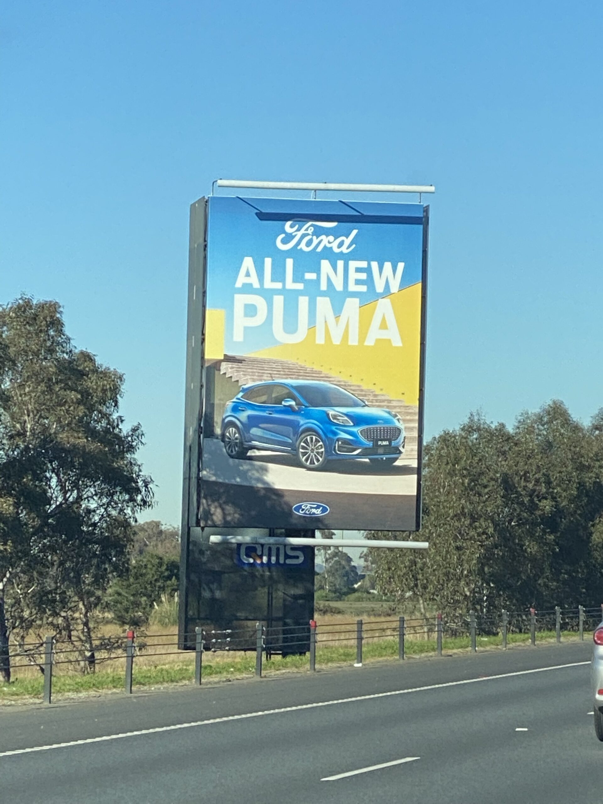 Large roadside billboard advertising Ford's all-new Puma with blue vehicle displayed against yellow and blue background, flanked by trees and rural fencing