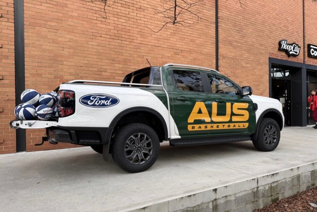 White and green Ford Ranger pickup truck with AUS Basketball livery parked against a brick building, displaying basketballs and sports equipment in the truck bed