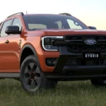 Orange Ford Ranger Hybrid pickup truck parked in grass field with lake and mountains in background at sunset
