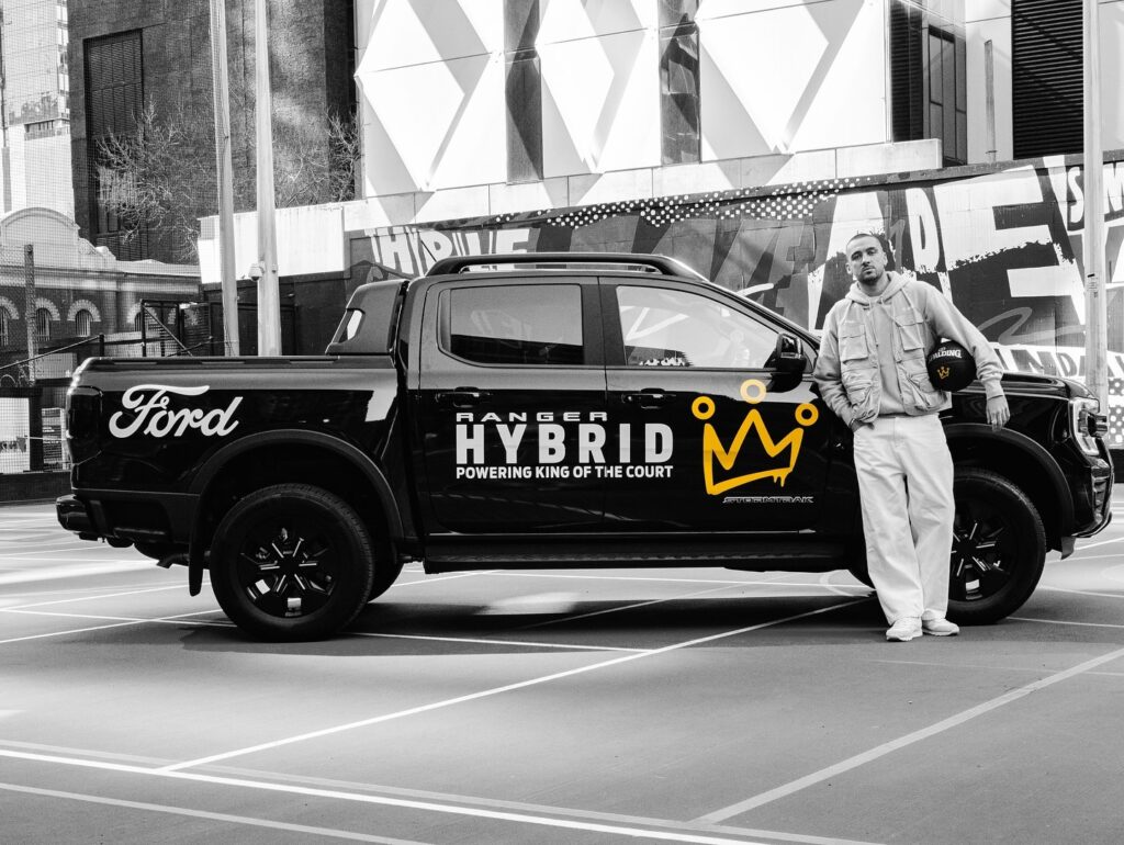 Man in white tracksuit standing beside black Ford Ranger Hybrid truck with yellow crown logo and 'Powering King of the Court' text on urban street with modern building backdrop
