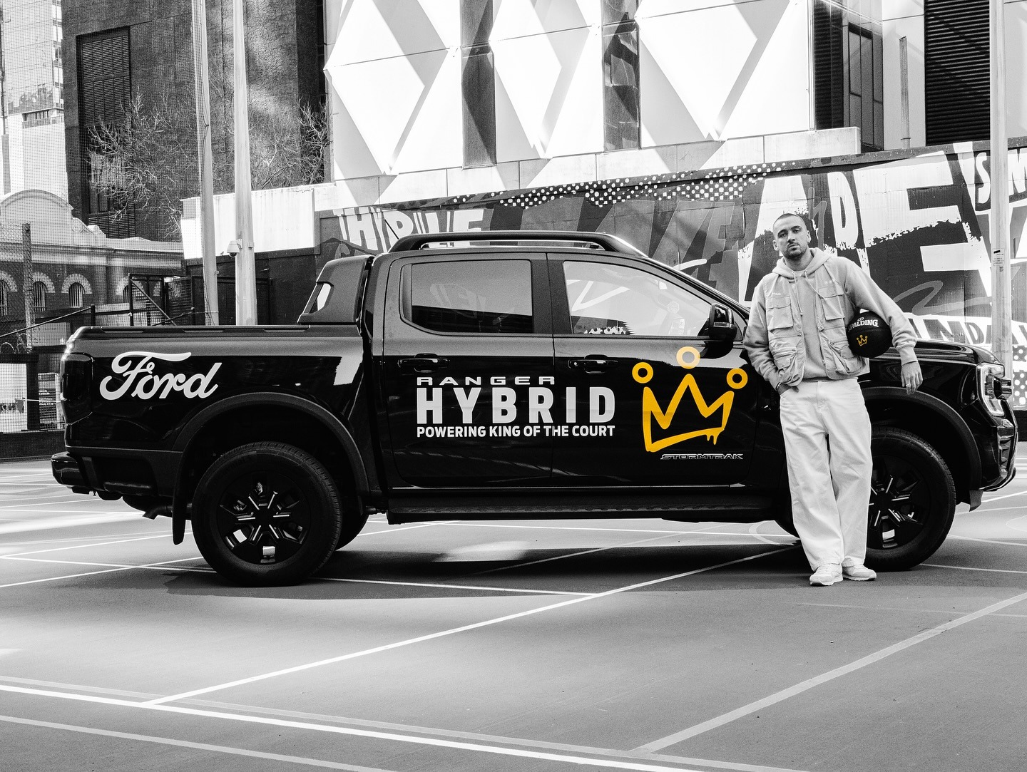 Man in white tracksuit standing beside black Ford Ranger Hybrid truck with yellow crown logo and 'Powering King of the Court' text on urban street with modern building backdrop