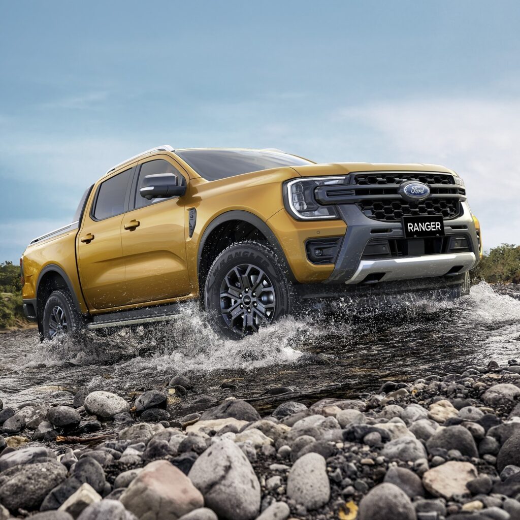 Gold Ford Ranger pickup truck fording through a rocky stream with water splashing around the wheels, clear sky in background