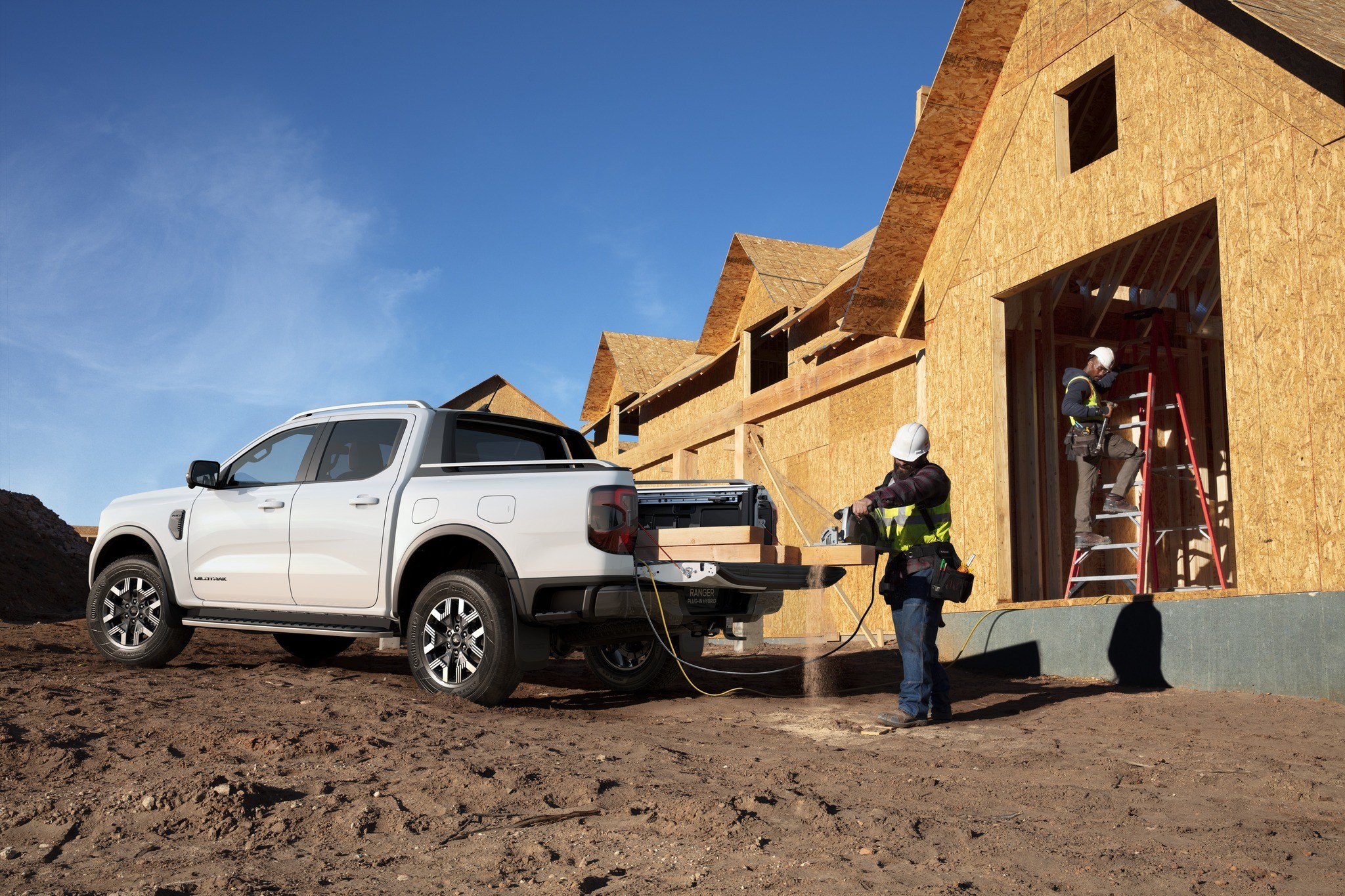 White Ford Ranger PHEV truck with ProPower Onboard system powering construction site, worker operating equipment from truck bed while others work on wooden building frame