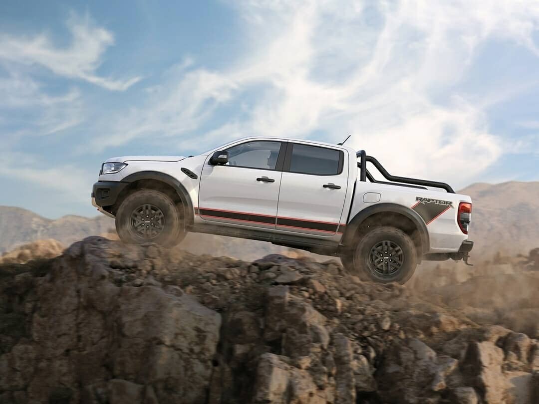 White Ford Ranger Raptor pickup truck positioned on rocky terrain with desert mountains in background under blue sky with clouds